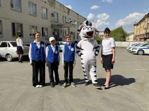 На изображении пять человек: четверо в синих жилетах и один в костюме зебры. Они стоят на парковке возле зданий.