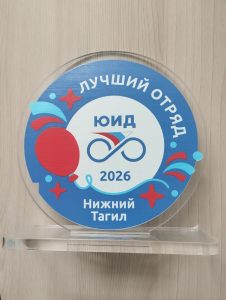 На изображении круглый знак с надписью «Лучший отряд ЮИД 2026 Нижний Тагил». Знак стоит на прозрачной подставке.
